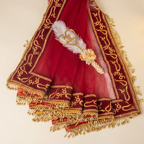 A deep red and gold embroidered dupatta with tassels, featuring a decorative white feather and gold heart brooch, laid out on a light beige background.